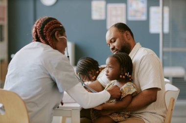 Hemşire beyaz üniformalı bir çocuk babası tarafından tutulan steteskoplu bir çocuğu muayene ediyor.