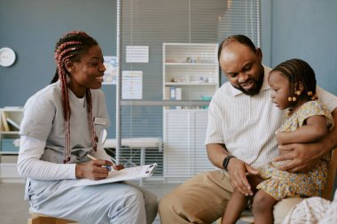 Hemşire önlüğü giymiş, babasıyla konuşan, modern klinikte kucağında küçük bir çocuk olan hemşire mükemmel ışıklandırma, dostça etkileşimi ve misafirperver çevreyi vurguluyor.