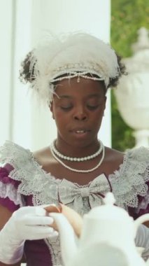 Klasik elbiseli ve tüylü başlıklı genç Afro-Amerikan bayanın dikey fotoğrafı. Açık terasta çay saatinde okuduğu kâğıtları döküyor.