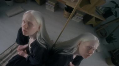 Halının üzerinde oturan, aynaya bakan ve oturma odasına bakan albino kızın yüksek açılı görüntüsü.