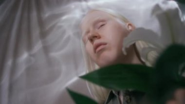 Yakından bakınca albino kızın yarı açık gözleri beyaz zambakların dalını büküyor ve çiçekleri kokluyor.