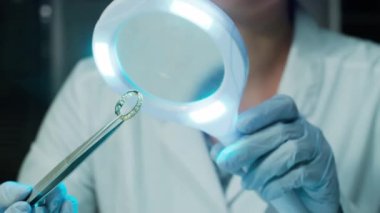 Kadın adli tıp uzmanının ya da mücevher eksperi, elinde cımbızla elmas yüzük tutan ve laboratuardaki aydınlatılmış büyüteç altında inceleyen eldivenli ellerine yakından bakın.