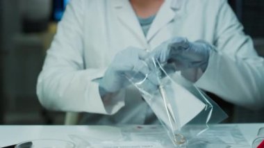 Beyaz önlüklü ve eldivenli kadın adli tıp uzmanı kanıt torbasından cımbızla yüzük çıkartıyor ve suç laboratuarında çalışırken ışıklı büyüteçle inceliyor.