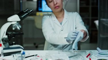 Kadın adli tıp uzmanının tek kullanımlık eldivenlerini giyip suç laboratuarında çalışırken aydınlatılmış büyüteç altındaki kanıtları incelerken görüntüsünü kaldır.