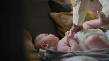 Sarı tişörtlü, beyaz tenli genç annenin yakın çekimi. Salondaki gri vagona uzanmış ağlayan ve kıpırdayan bir bebeğe bebek bezi takıyor.