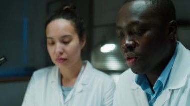 Siyah adli tıp uzmanı çantalarda toplanan kanıtları inceliyor ve yanında oturan ve notlar yazan kadın laboratuar teknisyeniyle konuşuyor.