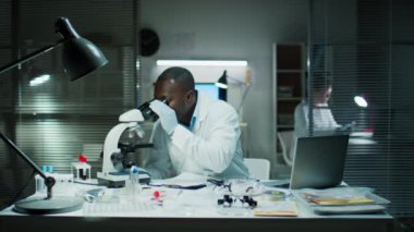 Afrikalı Amerikalı adli tıp teknisyeni suç laboratuarında çalışırken kanıtları mikroskop altında inceliyor ve bilgisayarda daktilo kullanıyor.