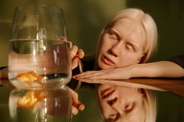 Medium closeup conceptual portrait of young woman with albinism posing for camera at table with mirror on it, watching pet goldfish swimming in small aquarium clipart