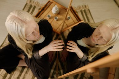 Yukarıdan bakıldığında, albinizmli genç bir kadın aynadaki yansımaya dokunarak kendini beğenmişlik ve kendini beğenmişliği ifade ediyor.