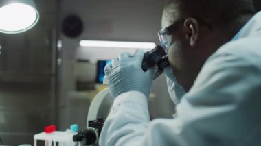 Eldivenli Afrikalı Amerikalı erkek serolog mikroskop altında kan örneğini inceliyor ve adli tıp laboratuarında çalışırken dijital tabletteki büyütülmüş resmi inceliyor.