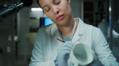 Tıbbi eldivenli ve beyaz önlüklü kadın adli tıp teknisyeni suç laboratuarında masasında oturuyor ve fırçayla bardağa parmak izi tozu sürüyor.