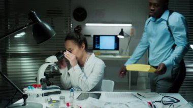 Suç laboratuarında mikroskopla çalışan kadın adli tıp uzmanı, sonra da dijital tablet üzerindeki kanıt analizinin sonuçlarını siyah erkek dedektife açıklıyor.