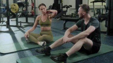 Spor giyimli genç bir adam ve kadın paspaslar üzerinde dinleniyor, spor salonunda antrenmandan sonra gülümsüyor ve sohbet ediyorlar.