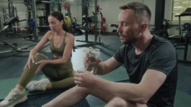 Spor kıyafetli yorgun bir adam spor salonunda mindere oturmuş idmandan sonra dinlenirken biberondan su içiyor ve yanında bir kadın buzağıya masaj yapıyor.