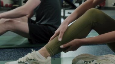 Beden eğitimi sonrası iyileşmek için baldır kaslarına masaj yapan, arka planda esneyen ve bağcıklarını bağlayan adamın yakın plan fotoğrafı.