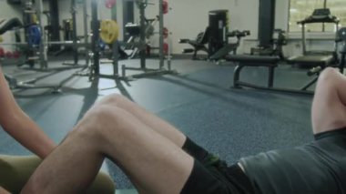 Spor kıyafetleri içinde kaslı bir adam spor salonunda egzersiz yaparken ayaklarını tutan kadın antrenörün yardımıyla mekik çekiyor.