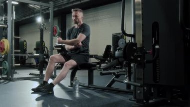 Spor kıyafetli bir adam spor salonunda bankta oturuyor, antrenmandan sonra dirseğini kontrol ediyor ve şişeden su içiyor.