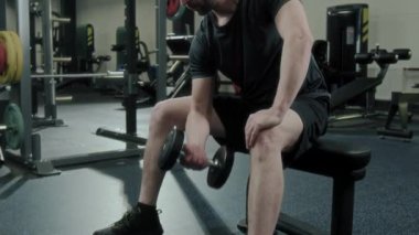 Spor kıyafetli bir adamın ağırlık kaldırma antrenmanı sırasında tek kollu halterde oturduğu ve tek kollu halterli kıvrımlar yaptığı pozlar.