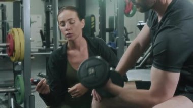 Kronometre kullanarak egzersiz yapan kadın jimnastik hocası, kişisel antrenman sırasında onu halterli buklelerle gezdirirken erkeklerle konuşuyor.