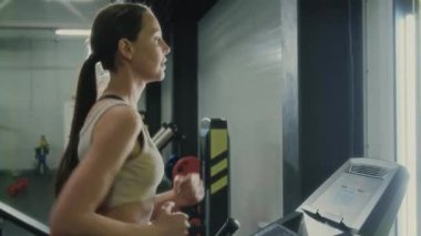 Spor giyimli genç bir kadının spor kıyafetleri içinde arka planda çeşitli fitness ekipmanlarıyla birlikte koşu bandında koşarken kardiyo çalışması yaparken çekimini kaldır.