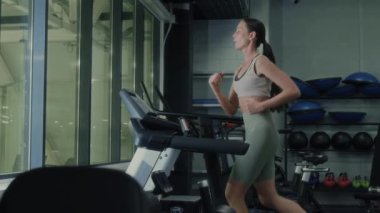 Spor kıyafetli genç bir kadın koşu bandı çalışmasını bitiriyor, su şişesini alıyor ve spor salonunu terk ediyor.