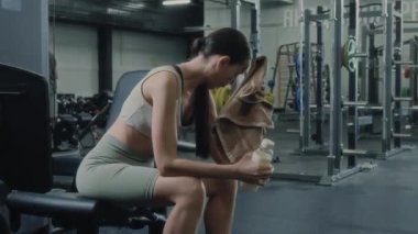 Spor kıyafetli genç bir kadın iyi donanımlı spor salonunda bankta oturuyor, alnından terini havluyla siliyor ve yüksek yoğunluklu idmandan sonra dinlenirken şişeden su içiyor.