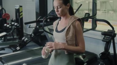Genç atletik kadın, omuzlarında havluyla modern spor salonunda duruyor ve egzersizden sonra dinlenirken şişeden su içiyor.