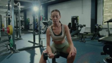 İyi donanımlı spor salonunda bankta oturan ve ağırlık antrenmanı sırasında tek kollu halterli kıvrımlar yapan genç bir kadın.