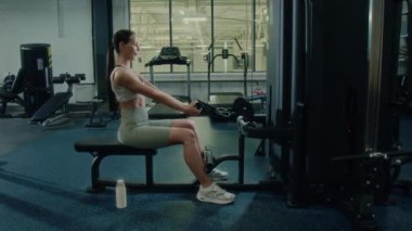 Genç atletik kadının kablolu yayında otururken, modern spor salonunda çeşitli egzersiz makineleriyle egzersiz yaparken görüntüsünü yakınlaştır.