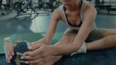 Spor kıyafetli genç bir kadın spor salonunda mindere oturuyor ve egzersizden sonra esneme hareketleri yaparken tek bacaklı bir viraj yapıyor.