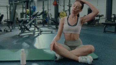 Sıska bir kadın spor salonunda bacak bacak üstüne atarak oturuyor ve antrenman sonrası omuzlarını ve boynunu esnetiyor.