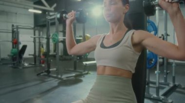 Spor salonunda egzersiz yaparken halterli omuz presi yapan genç bir kadın.