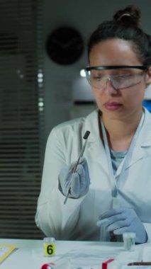 Eldivenli, laboratuvar önlüklü ve koruyucu gözlüklü kadın adli tıp uzmanının dikey görüntüsü balistik kanıtları cımbızla kaptan çıkarıp inceleme yaparken...