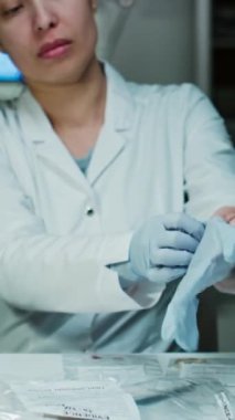 Laboratuvar önlüklü kadın adli tıp uzmanının tek kullanımlık eldivenler giyerken, ışıklı büyüteç alırken ve olay yerindeki kanıtlarla dolu kabı incelerken dikey bir fotoğrafı.