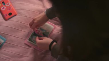 Tanımlanamayan kızın, idol kartını küçük pembe albüme koyarken, sayfayı çevirirken ve yatakta otururken başka bir resmi düzeltirken yakın plan görüntüsü.