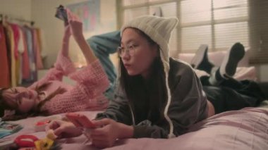 Beyaz şapkalı Koreli genç, elinde akıllı telefonuyla yatakta rahatlıyor, liçi yiyor ve beyaz kız arkadaşıyla k-pop müzik dinliyor.