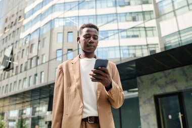 Açık havada duran ve akıllı telefondan müzik listesi seçen Afro-Amerikalı genç işadamının düşük açılı görüntüsü.