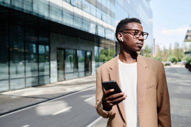 Z Gen yöneticisinin gözlüklü resmi giyip işe giderken müzik ya da podcast dinlerken orta boy görüntüsü.