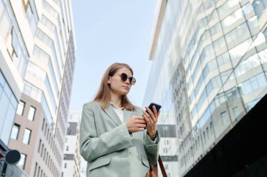 Beyaz tenli, güneş gözlüğü takan genç bir kadının iş yerinde dışarıda durup akıllı telefondan internette sörf yaptığı düşük açılı görüntü.