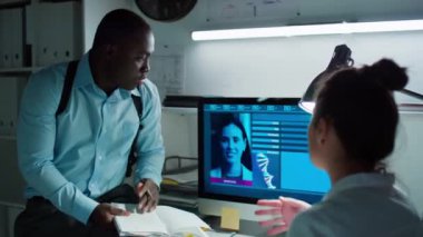 Afrika kökenli Amerikalı erkek araştırmacı bilgisayar ekranını işaret ediyor ve laboratuarda birlikte çalışırken kadın adli tıp uzmanıyla şüpheli profilini tartışıyor.