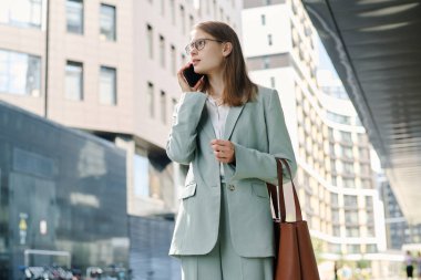 İş bölgesinde cadde boyunca yürürken telefonla konuşan beyaz bir kadının düşük açılı görüntüsü.