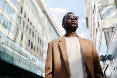 Orta açılı, genç, Afro-Amerikan erkek ofis çalışanı güneş gözlüğü takıyor ve iş bölgesinde yürüyor.