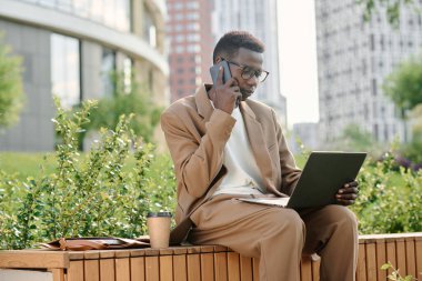 Genç nesil Afro-Amerikan adam bankta oturuyor telefonda müşteriyle konuşuyor ve bilgisayar ekranına bakıyor.