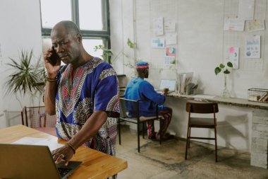 Afrikalı Amerikalı iş adamı, çalışanı arka planda finansal sunum yaparken telefonda iş ortağıyla konuşuyor.