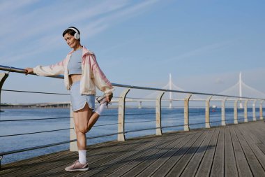 Pastel renkli spor giysiler giymiş, kıdemli beyaz kadın rıhtımda koşmadan önce diz eklemlerini ısıtıyor.