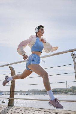 Rıhtımda hızlı koşan olgun bir kadının dikey çekimi. Kardiyo yaparken müzik dinliyor.