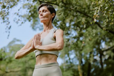 Düşük açılı çekimde, sportif emekli beyaz kadın yaz günü parkta yoga pozisyonunda dururken dinleniyor.