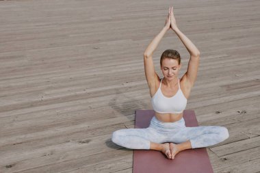 Genç ve güzel bir kadının pozisyonunu alıp meditasyon yaptığı yüksek açılı bir çekim. Açık gökyüzünün altında, ahşap zeminde tek başına yapıyor.