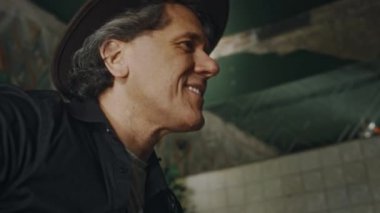 Orta yaşlı, gülümseyen, şapka takan, elektro gitar çalan, arka planda tanınmayan grup arkadaşlarıyla eğlenen beyaz erkek müzisyenlerin yakın çekimi.