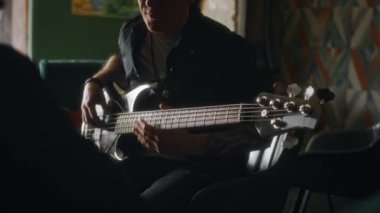 Orta yaşlı, gülümseyen, beyaz bir erkek müzisyenin grup arkadaşlarıyla oturup, boş zaman geçirip elektro gitar çalarken orta yavaş çekimi.